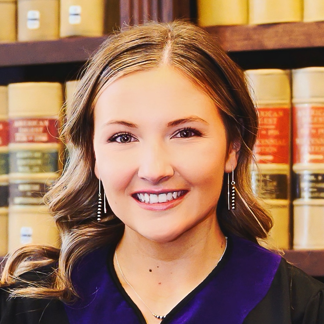 Woman in a black academic gown with a purple collar smiles in front of shelves filled with large legal books.